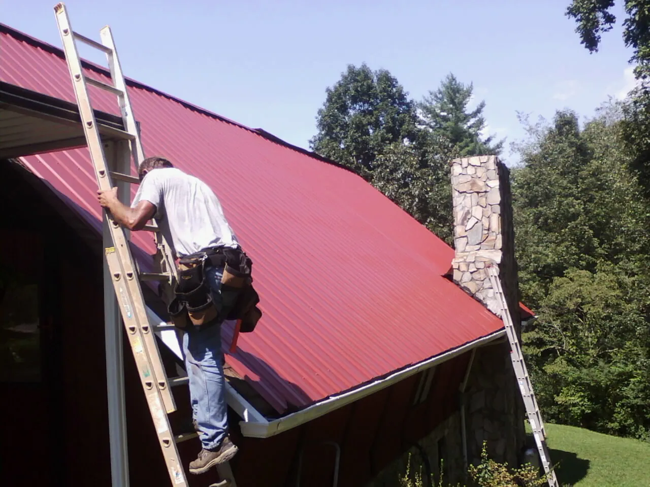 Roofer on a ladder beside a red metal roof and stone chimney — Soffit & Fascia Repair by Roofing Brothers in West Boylston